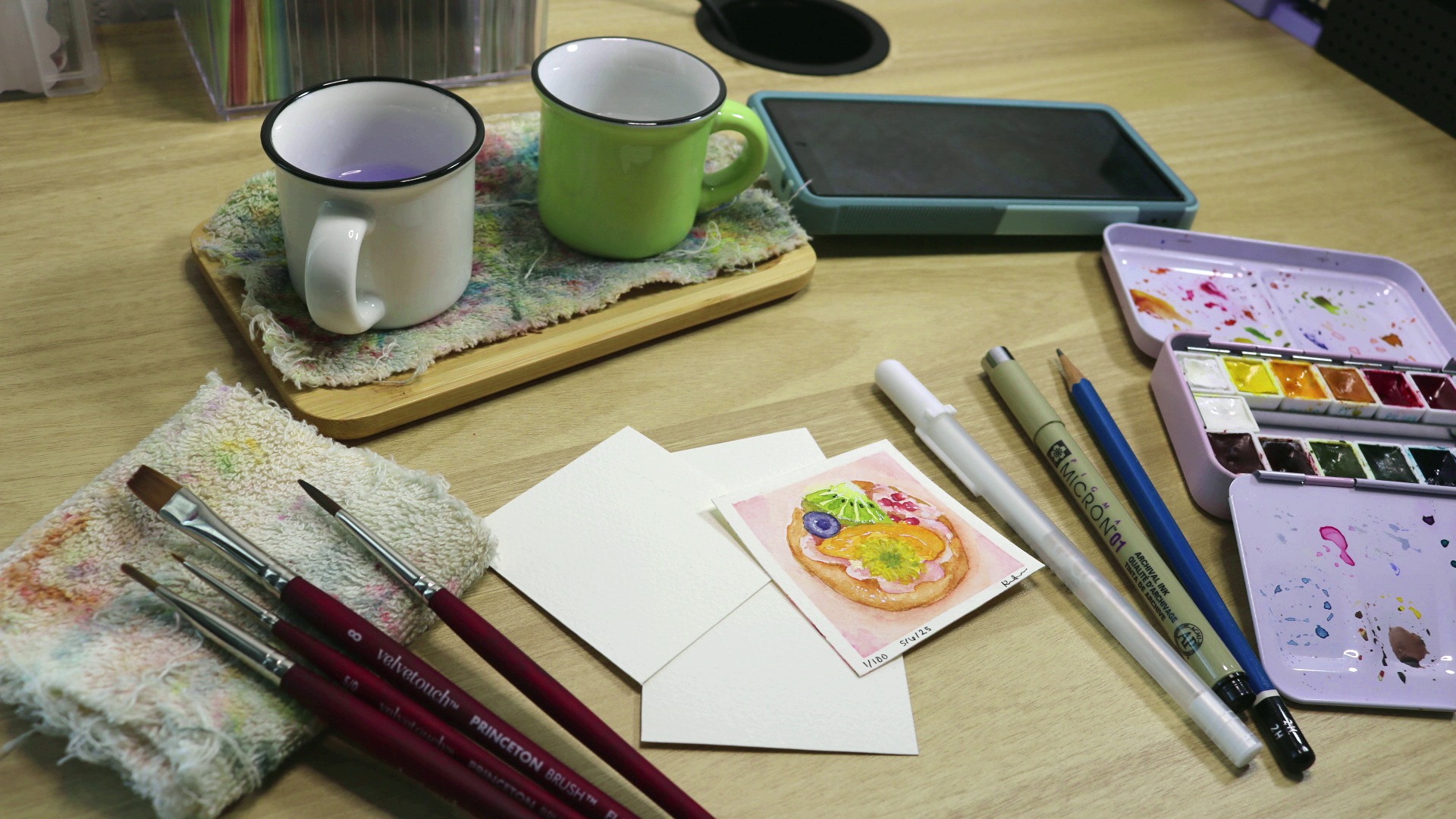 Everything needed for a painting challenge is arranged on a desk: small pieces of watercolor paper, water cups, a watercolor palette, paintbrushes and drawing utensils.