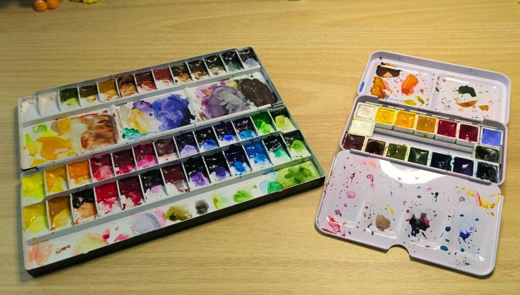 two watercolor palettes lay open next to each other on a desk